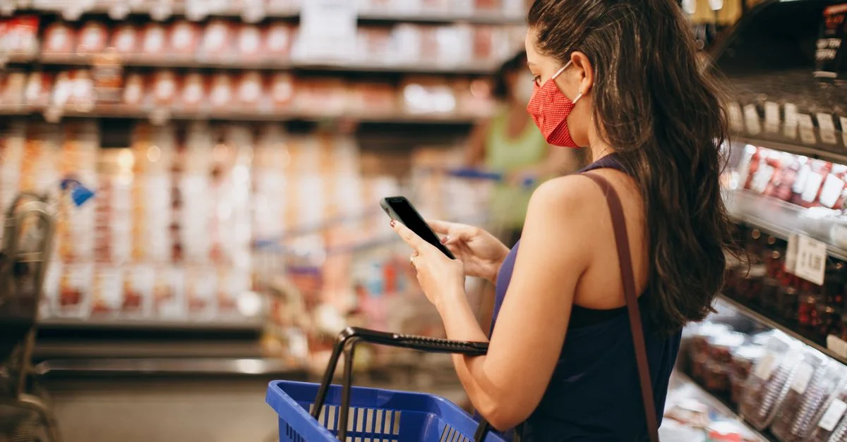 Une femme avec un masque fait ses courses dans un supermarché en utilisant son smartphone et un panier à la main.