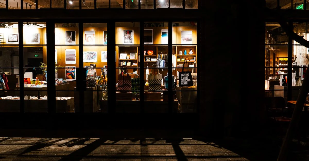 Une vue nocturne d'une vitrine de magasin présentant des sacs et des affiches, créant une atmosphère urbaine chaleureuse.