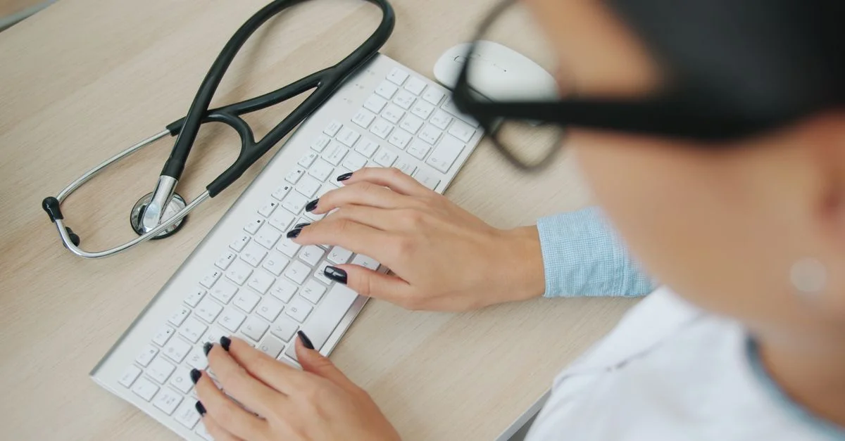 Le médecin utilise un clavier d'ordinateur ; un stéthoscope se trouve à proximité, sur le bureau.